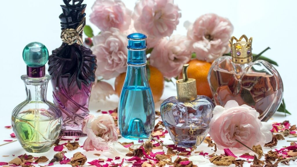 A imagem apresenta seis frascos de perfume de formatos diferentes, com rótulos coloridos, dispostos sobre uma superfície branca. Os frascos estão cheios de diversas fragrâncias.
