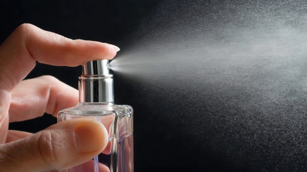 Uma pessoa borrifa um frasco transparente de perfume contra um fundo preto.
