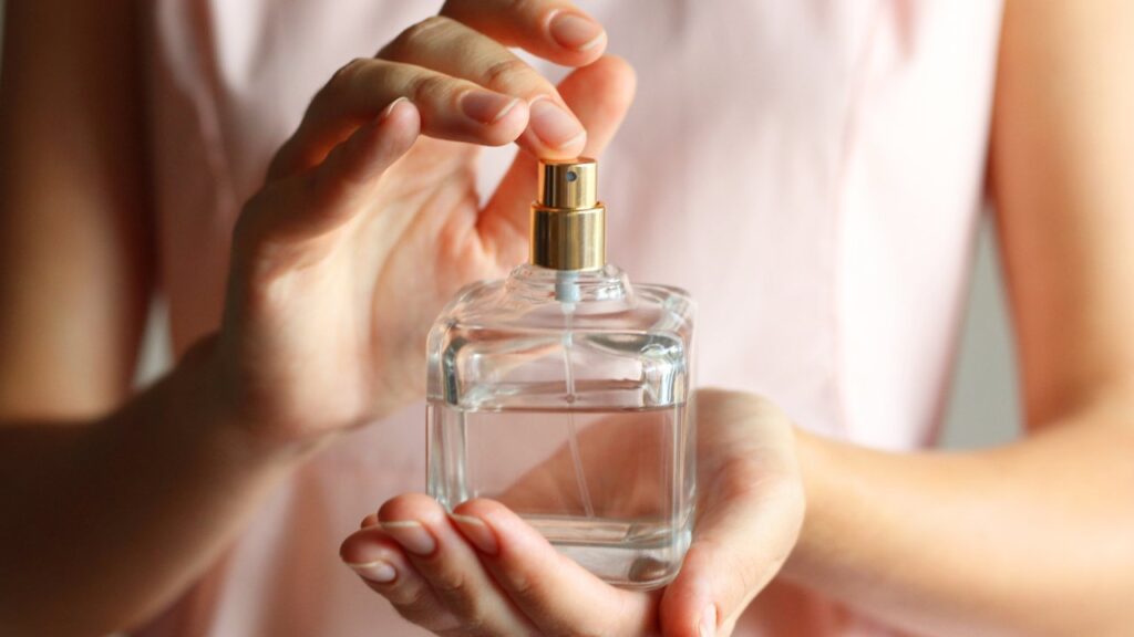 Uma mulher de camisa rosa segura um frasco transparente de perfume nas mãos.
